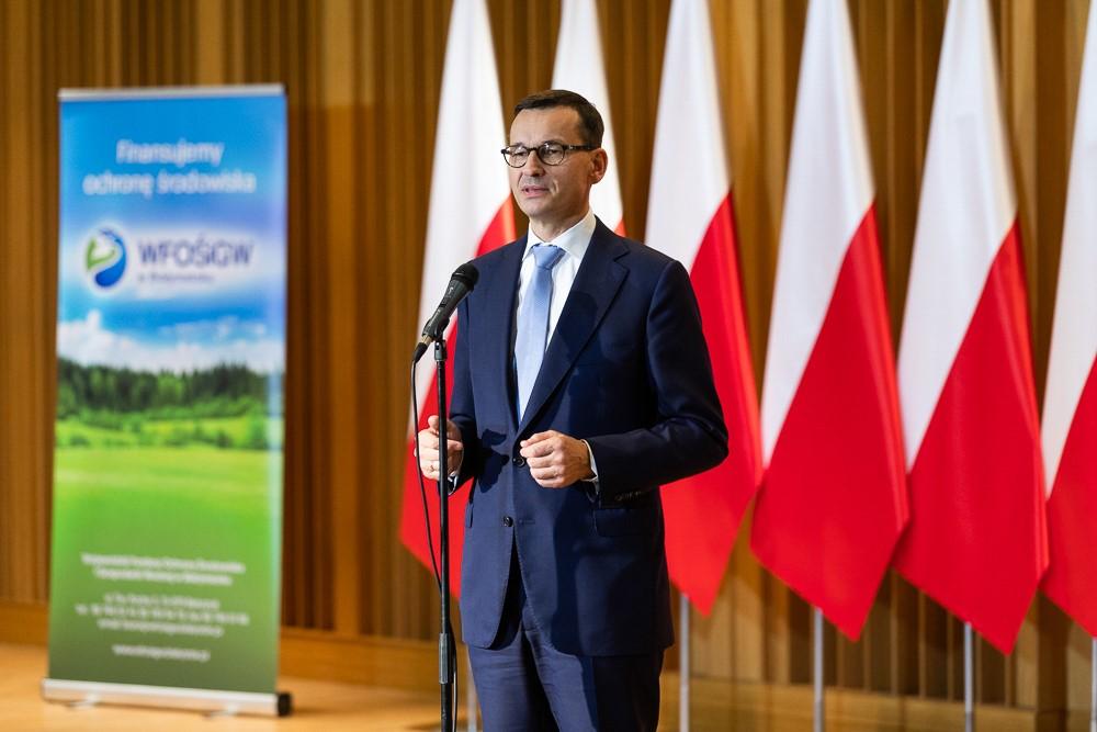 Premier Mateusz Morawiecki stoi przy mikrofonie na tle flag.