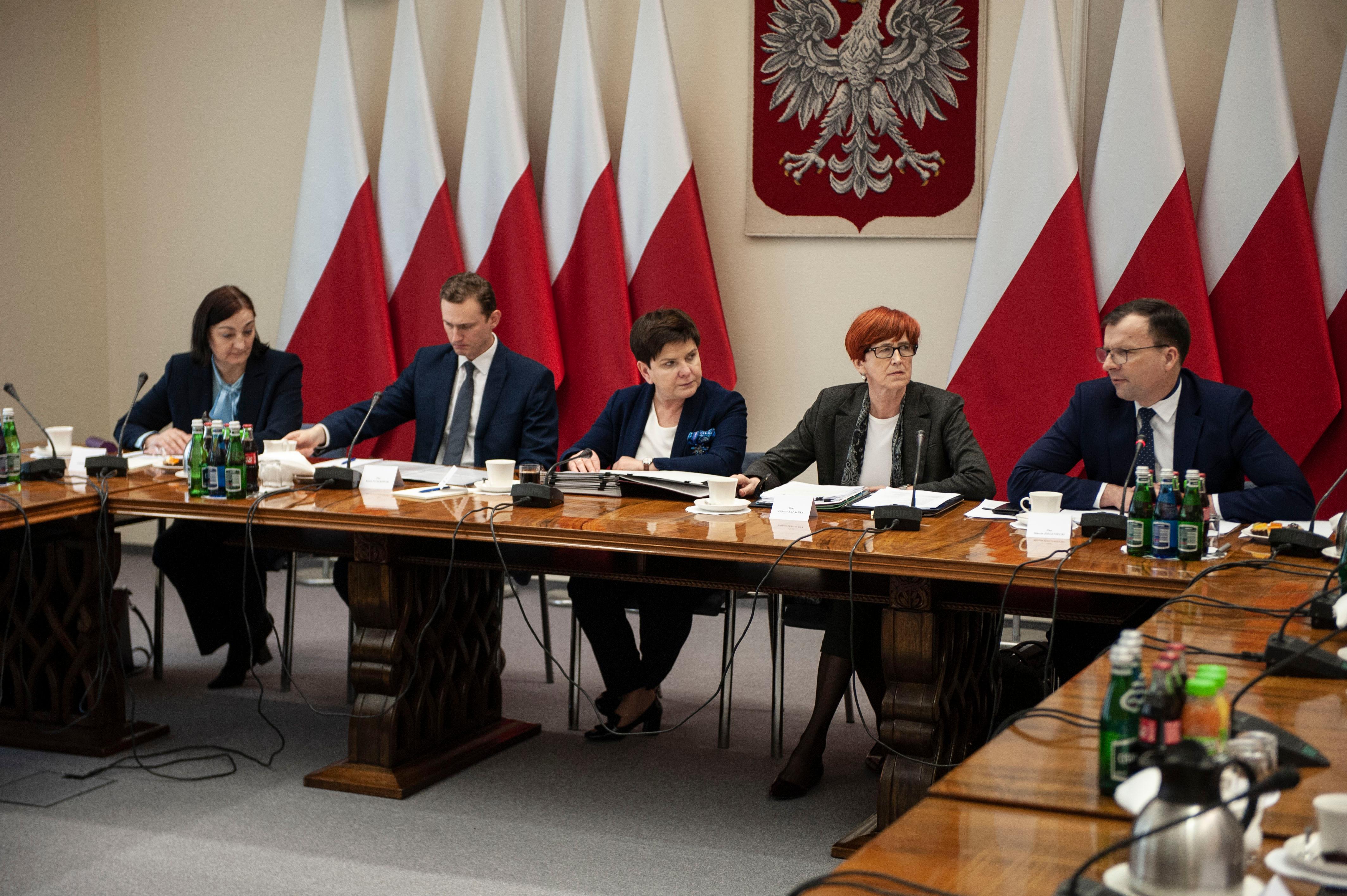 Wicepremier Beata Szydło ze współpracownikami siedzi na tle godła i biało-czerwonych flag.