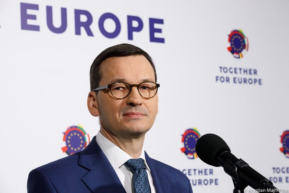 Premier Mateusz Morawiecki stoi przed mikrofonem. 