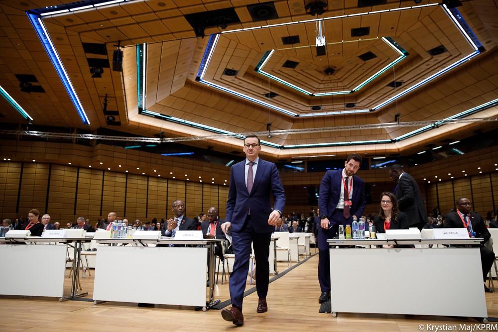 Premier Mateusz Morawiecki idzie, a w tle siedzą uczestnicy forum.