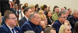 Widok z boku na salę, na krzesłach w rzędach siedzą ludzie, pomiędzy rzędami krzeseł stoi duża kamera na statywie, w tle żółte ściany.
