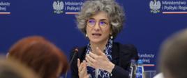 Przedstawicielka Komisji Europejskiej siedzi w środku przy stole i mówi do mikrofonu. 