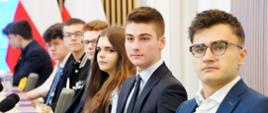 Widok z boku na prawą stronę sali, przy ustawionych w rząd stolikach siedzą młodzi ludzie, przed nimi mikrofony.