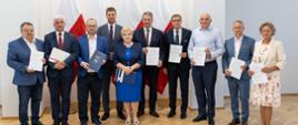 Zdjęcie zbiorowe, grupa ludzi stoi pod ścianą, pokazują trzymane w rękach dokumenty.