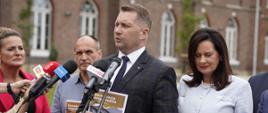 Na dworze przed szaro-brązowym budynkiem stoi minister Czarnek, trzyma w ręku broszurkę w brązowej okładce i mówi do mikrofonów na stojakach, obok niego stoi kobieta w białym ubraniu i trzyma taką samą broszurkę, za nimi kilka osób.
