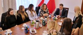 Widok z góry pod kątem, po obu stronach długiego prostokątnego stołu siedzi kilkanaście młodych osób, u szczytu stołu minister Czarnek, za nim dwie polskie flagi i biała ściana, po lewej zasłonięte firankami okna.