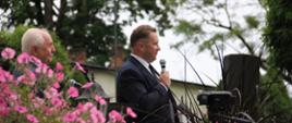 Na dworze pod drzewami minister Czarnek stoi i mówi do mikrofonu, na pierwszym planie różowe kwiaty.