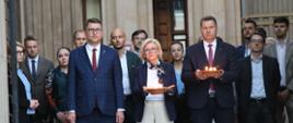 Przed wejściem do szarego budynku stoi grupa ludzi, z przodu minister Czarnek i wiceminister Machałek trzymają w rękach płonące znicze.