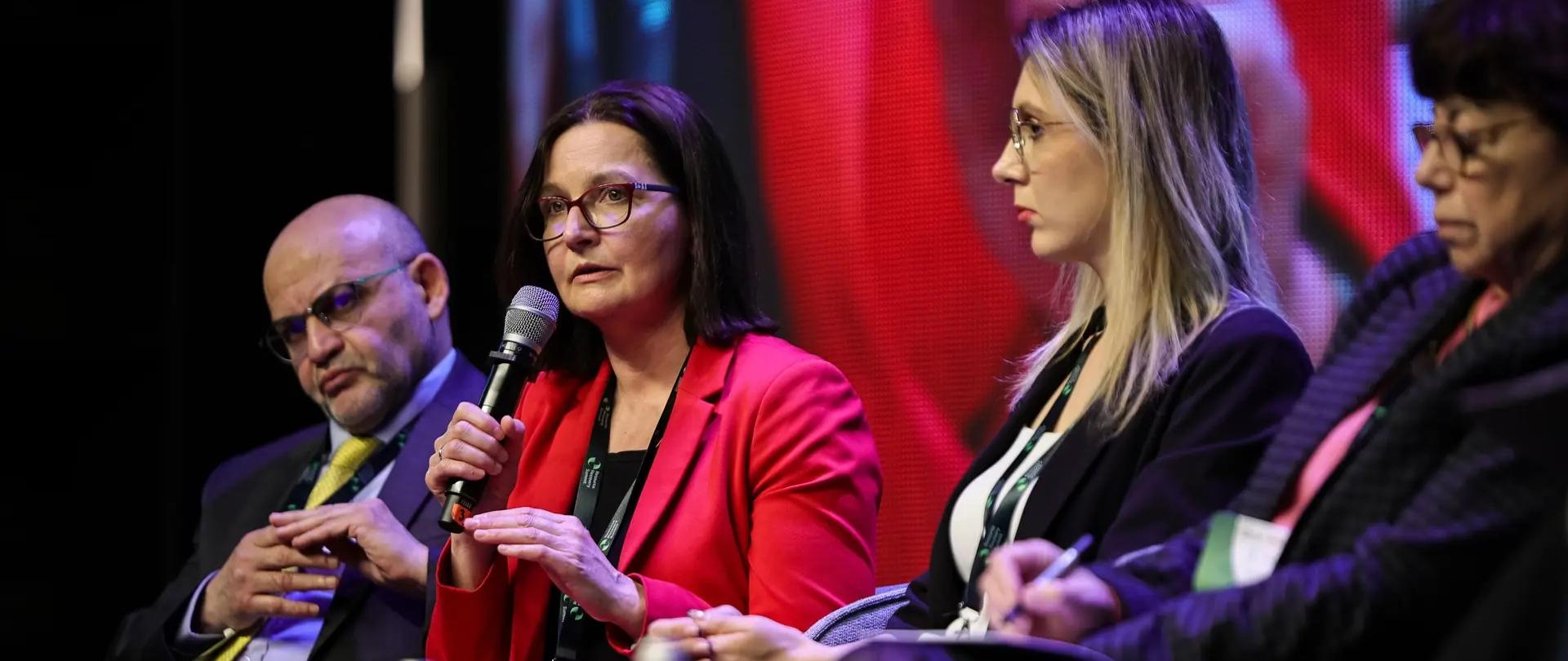 Wiceministra klimatu i środowiska Anita Sowińska ubrana w czerwony garnitur siedzi na scenie wśród czterech osób (pozostali ubrani w ciemne garnitury i garsonki). 