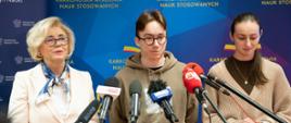 Wiceminister Machałek w białym ubraniu stoi na tle niebieskiej ścianki z napisem Karkonoska Akademia Nauk Stosowanych, obok niej młody chłopak w szaro-brązowej bluzie z kapturem i dziewczyna w sweterku, przed nimi kilka mikrofonów na stojakach.