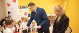 inister podaje rękę małemu chłopcu, za nim stoi ubrana na czarno kobieta, dalej grupa dzieci, za nimi żółta ściana.