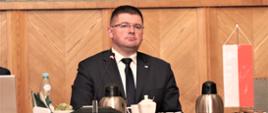 Mężczyzna siedzi za stołem obok flaga Polski, na stole filiżanki, woda, poczęstunek