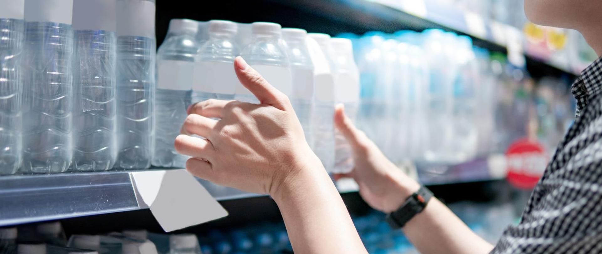 Na zdjęciu widoczne są półki, na których stoją plastikowe butelki. Dodatkowo widać dłonie, które wstawiają na półkę zgrzewkę z napojami.