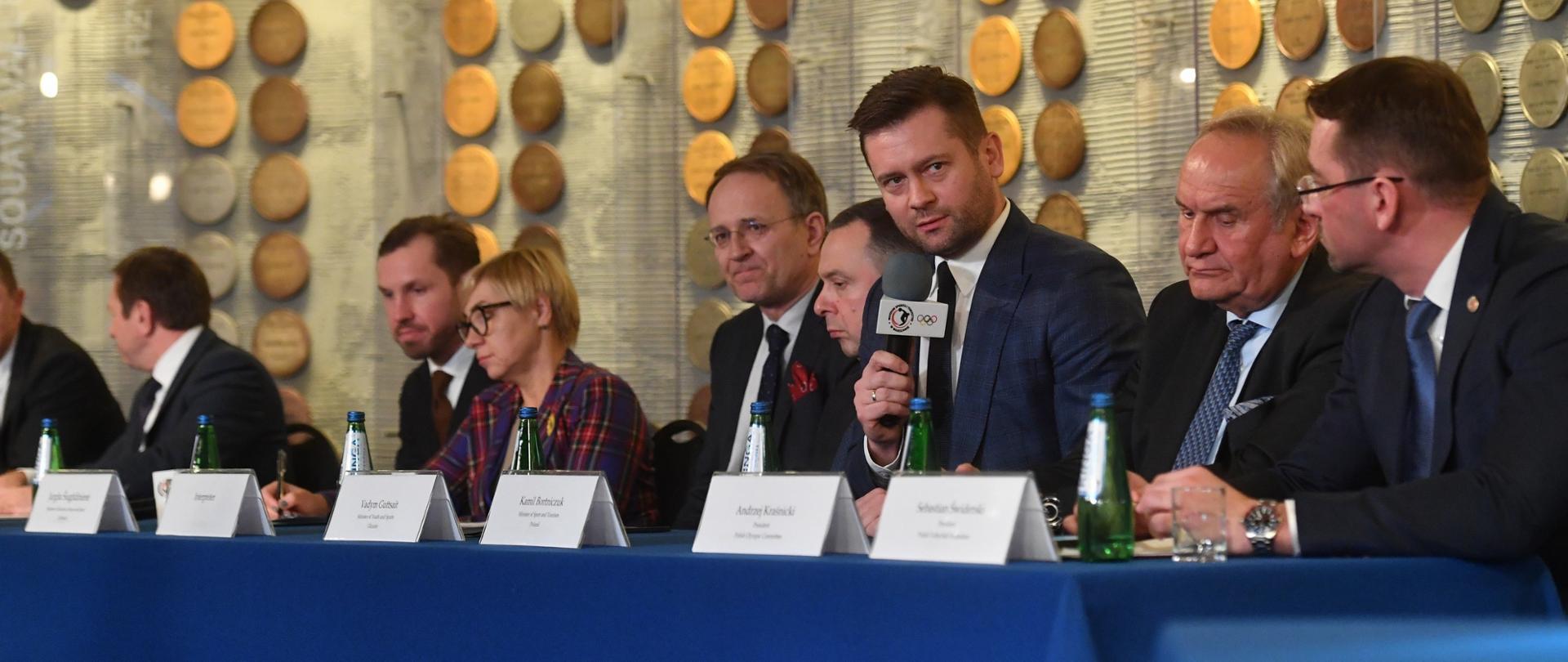 Prezesi polskich związków sportowych podpisali wspólne stanowisko w sprawie utrzymania sankcji sportowych nałożonych na Rosję i Białoruś. Za stołem siedzą przedstawiciele delegacji rządowych Polski, Ukrainy, Litwy, Czech i Słowacji. Minister Kamil Bortniczuk przemawia do mikrofonu