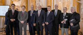 W eleganckiej sali ze zdobionymi złoceniami ścianami na podłodze z żółtych i brązowych czworokątnych płyt stoi szereg ośmiu mężczyzn w garniturach.