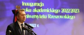 Minister Czarnek stoi przy mównicy i mówi do mikrofonu, za nim ściana z napisem Inauguracja roku akademickiego.
