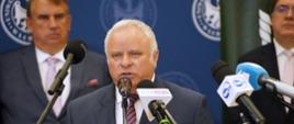 Mężczyzna w garniturze mówi do kilku mikrofonów na stojakach, za nim stoi dwóch mężczyzn.
