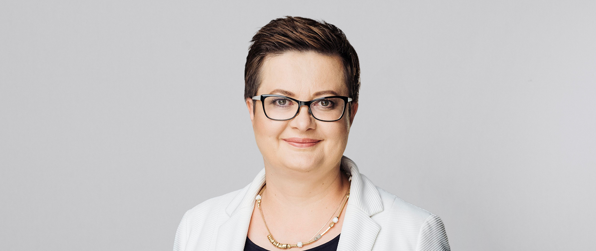 Na tle jasnoszarej ściany stoi wiceminister Katarzyna Lubnauer w białej marynarce.