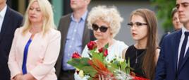 Trzy elegancko ubrane kobiety stoją na dworze, jedna trzyma bukiet białych i czerwonych kwiatów.