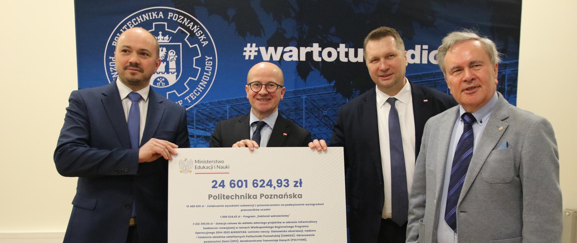 Na tle granatowej ścianki stoi minister Czarnek w towarzystwie trzech mężczyzn w garniturach, dwóch z nich trzyma wielki symboliczny czek z napisem 24 601 624,93 zł.