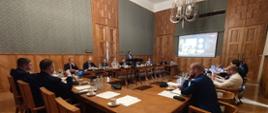 Spotkanie inaugurujące działalność Zespołu do spraw wsparcia młodzieżowych drużyn pożarniczych, uczestnicy spotkanie siedzą przy stole konferencyjnym, z tyłu ekran z telekonferencją.