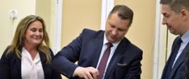 Minister Czarnek stoi i trzyma w reku metalowy cylinder, obok niego stoi kobieta w czarnym swetrze i mężczyzna w garniturze, za nimi żółte ściany.