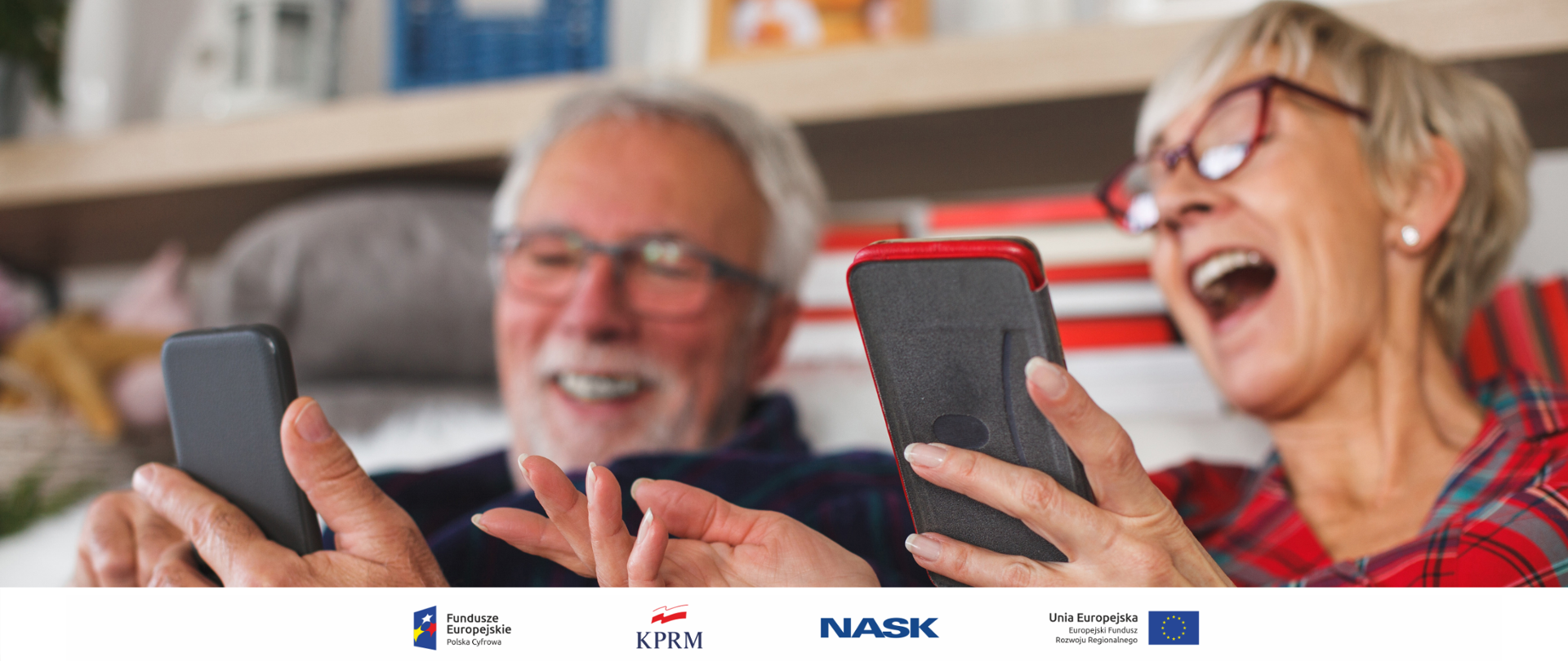 Dwoje seniorów (kobieta i mężczyzna) siedzą na kanapie z telefonami w rękach. Oglądają coś zabawnego - oboje się śmieją pokazując sobie ekranu urządzeń.