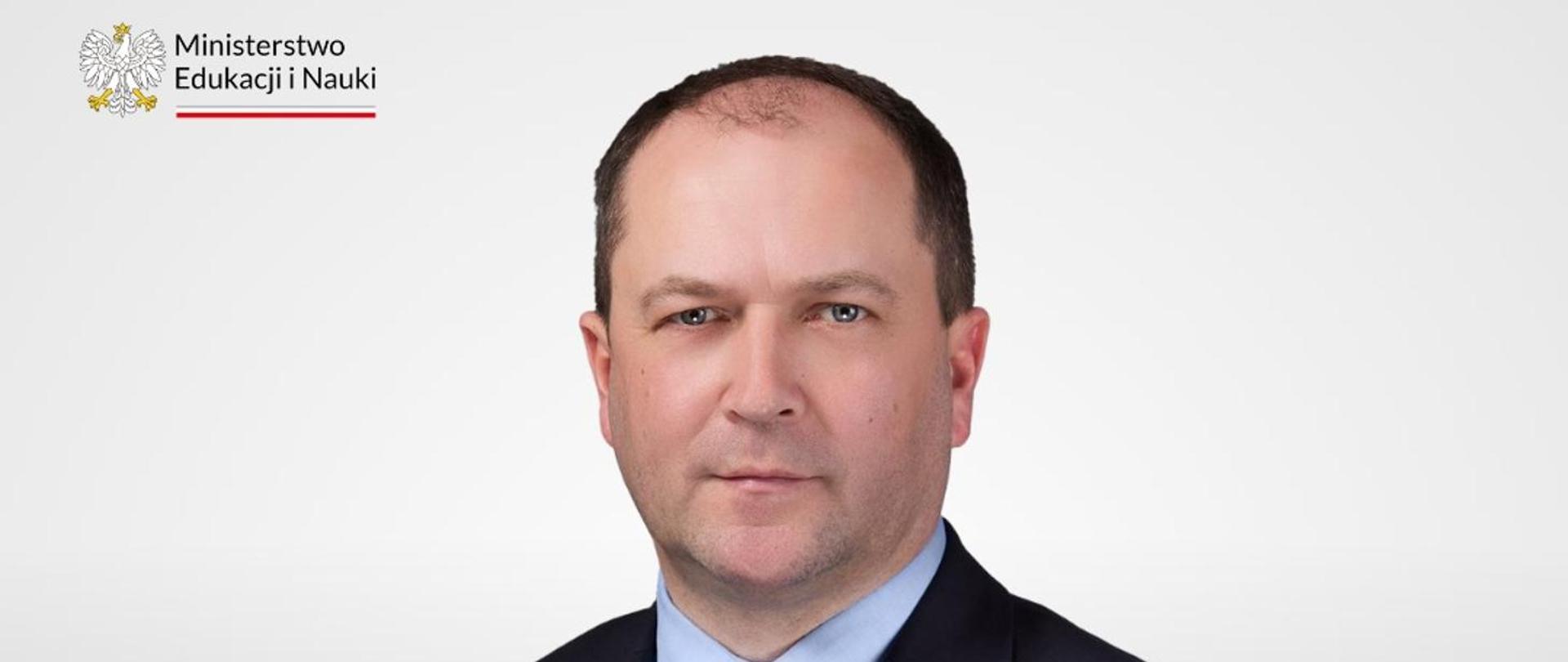 Na tle jasnoszarej ściany stoi wiceminister Kiepura w czarnym garniturze.