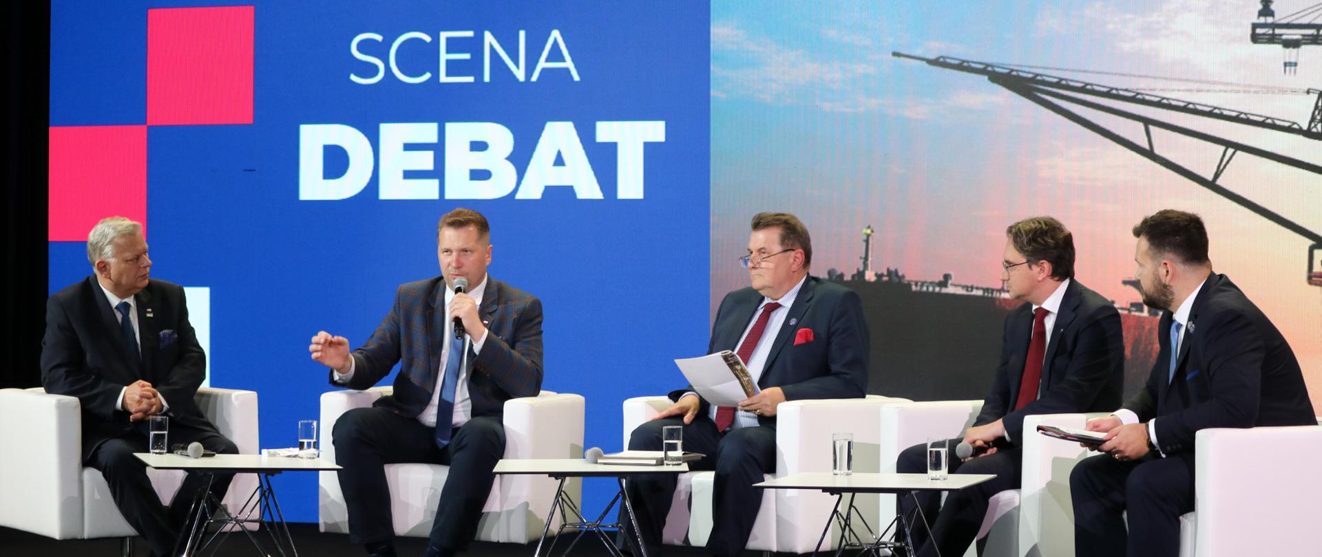 Uczestnicy debaty siedzą na krzesłach, za nimi ekran z napisem Scena debat.