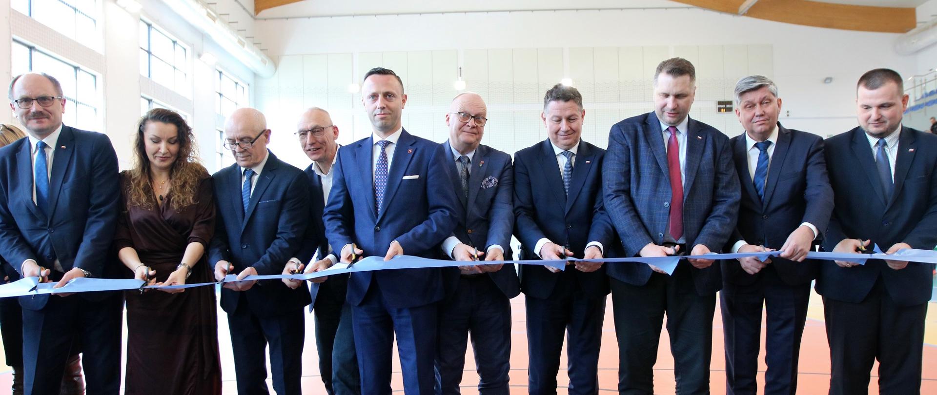 Na środku hali sportowej stoi szereg elegancko ubranych ludzi, każdy trzyma nożyczki, wszyscy przecinają biegnącą w poprzek niebieską wstęgę.