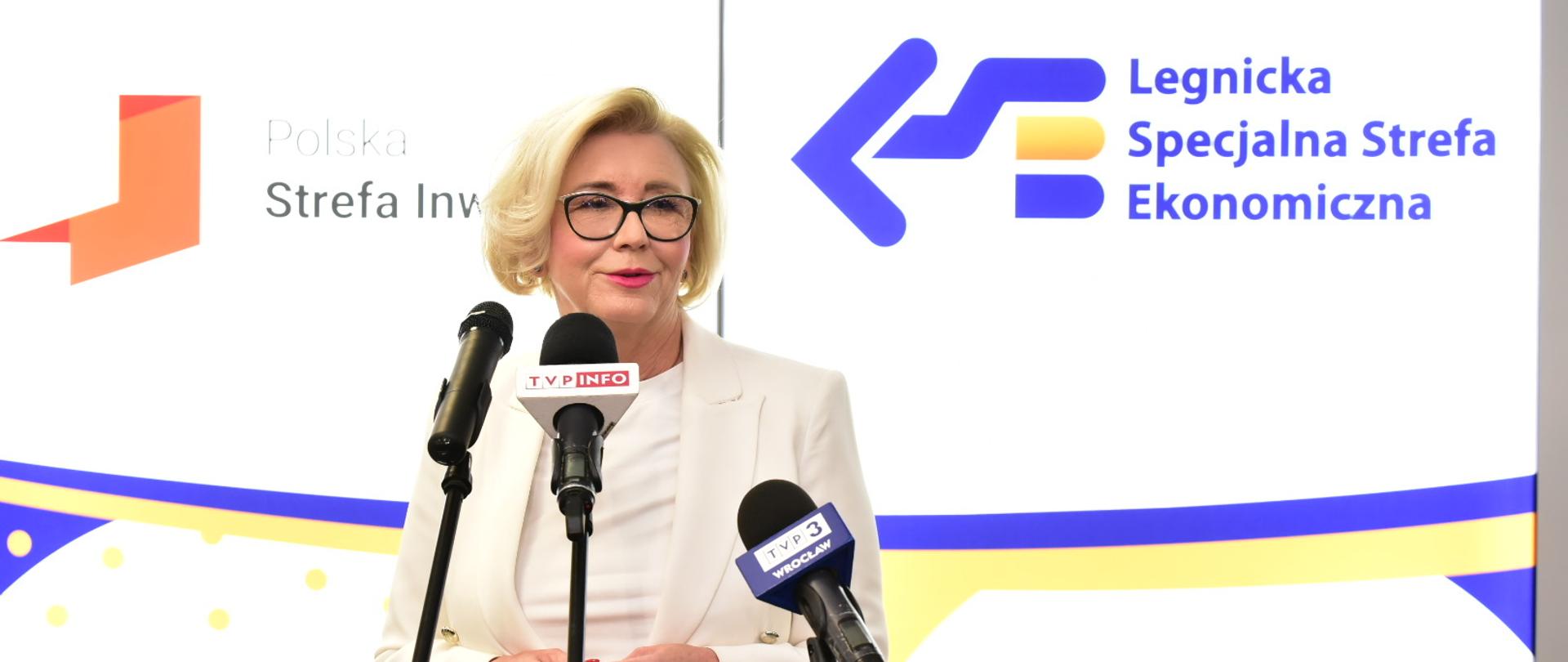 Wiceminister Machałek w białym ubraniu stoi i mówi do trzech mikrofonów na stojakach, za nią biała ściana z napisem Legnicka Specjalna Strefa Ekonomiczna.