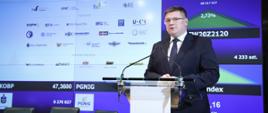Dzień Edukacji Finansowej z udziałem wiceministra Tomasza Rzymkowskiego, wiceminister Tomasz Rzymkowski przemawia, stoi przed mównicą, za nim wyświetlana jest prezentacja.