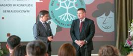 Przed siedzącymi na krzesłach ludźmi stoi mężczyzna w garniturze i wiceminister Rzymkowski który mówi do mikrofonu na stojaku, za nimi ekran z napisem Bez korzeni nie zakwitniesz. Moja Wielka i Mała Ojczyzna.