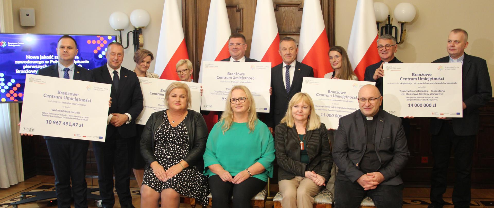 Grupa osób. Na pierwszym planie trzy kobiety i mężczyzna siedzą na krzesłach za nimi stoi grupa kobiet i mężczyzn trzymają w rękach symboliczne czeki. W tle biało-czerwone flagi.