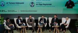 Sześć osób, czterech mężczyzn i dwie kobiety, siedzą w jasnych fotelach na scenie. Wszyscy są ubrani w formalne stroje. Między nimi stoją małe, drewniane stoliki z butelkami wody i szklankami. W tle widoczny jest duży ekran, na którym wyświetlane są informacje o panelistach.