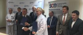 Grupa ludzi stoi za białym stołem, z przodu mężczyzna w garniturze i mężczyzna w białej koszuli podają sobie ręce, każdy trzyma niebieską teczkę.