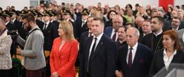 Widok od przodu na wypełnioną stojącymi ludźmi salę, w pierwszym rzędzie minister Czarnek i kobieta w pomarańczowym garniturze.