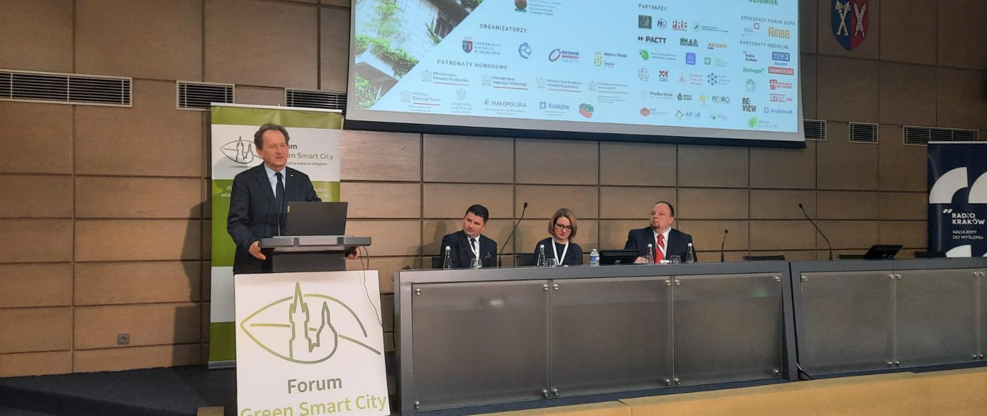 Wiceminister Bernacki stoi za mównicą z zielonym napisem Forum Green Smart City na białym tle, za nim ściana wyłożona drewnianymi panelami, obok siedzą trzy osoby.
