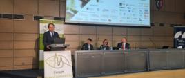 Wiceminister Bernacki stoi za mównicą z zielonym napisem Forum Green Smart City na białym tle, za nim ściana wyłożona drewnianymi panelami, obok siedzą trzy osoby.