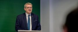 Wiceminister Murdzek stoi za mównicą i mówi do mikrofonu, za nim zielona tablica.