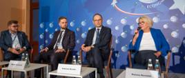 Wiceminister Machałek i trzech mężczyzn w garniturach siedzą na krzesłach, za nimi niebieska ściana, Minister Machałek mówi do trzymanego w ręku mikrofonu.