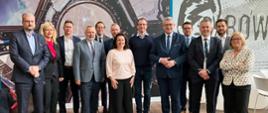 Na tle ściany w szare wzorki stoi duża grupa ludzi, pośrodku wiceminister Murdzek.