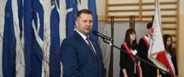 Minister Czarnek stoi i mówi do mikrofonu na stojaku, dalej biało-czerwony sztandar i troje młodych ludzi przepasanych biało-czerwonymi wstęgami.