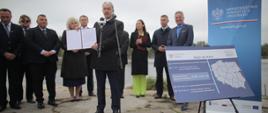 przed mikrofonem Jerzy Kwieciński, który trzyma otwarty dokument z opinią, a za nim uczestnicy uroczystości