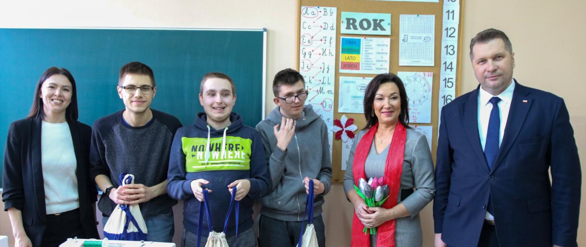 Minister Czarnek, kobieta w szarej sukience z czerwonym szalikiem i czworo młodych ludzi uśmiechają się stojąc pod żółtą ścianą, na której wisi zielona szkolna tablica i korkowa tablica z przyczepionymi kolorowymi kartkami.
