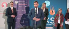 Minister Kamil Bortniczuk przemawia do zebranych podczas konferencji prasowej na temat warsztatów o przeciwdziałaniu przemocy w sporcie. W tle wiceminister Marcin Romanowski z Ministerstwa Sprawiedliwości oraz przedstawiciele Instytutu Sportu i Światowej Organizacji Zdrowia.