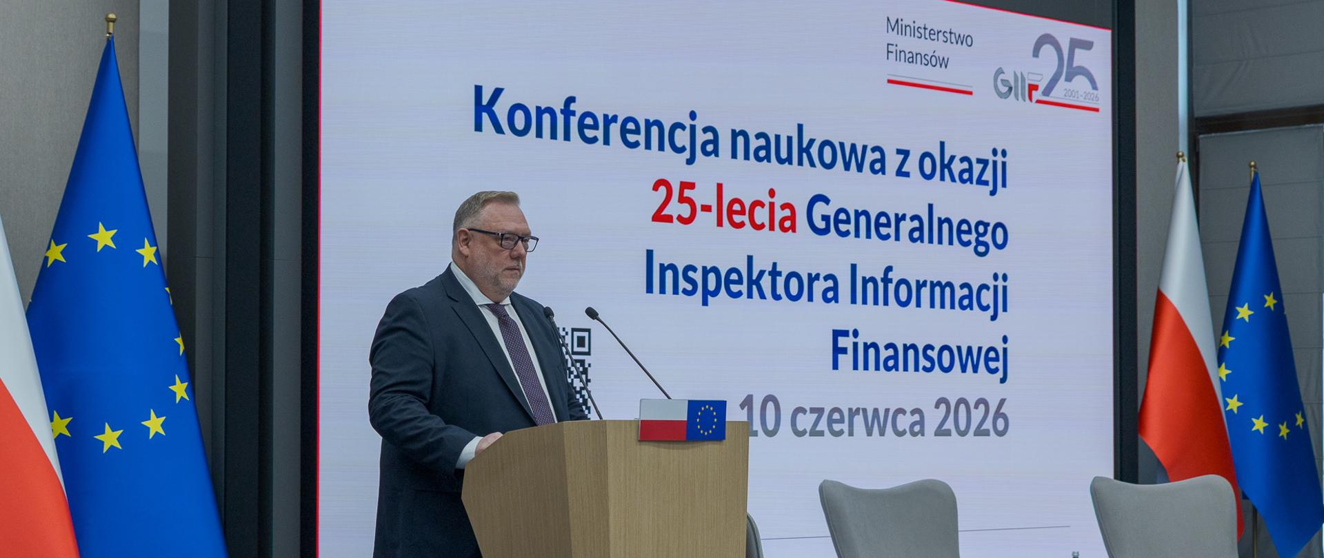 Generalny Inspektor Informacji Finansowej Jarosław Żółtowski stoi przy mównicy podczas konferencji naukowej Ministerstwa Finansów z okazji 25-lecia Generalnego Inspektora Informacji Finansowej, na tle ekranu z tekstem oraz flag Polski i Unii Europejskiej.