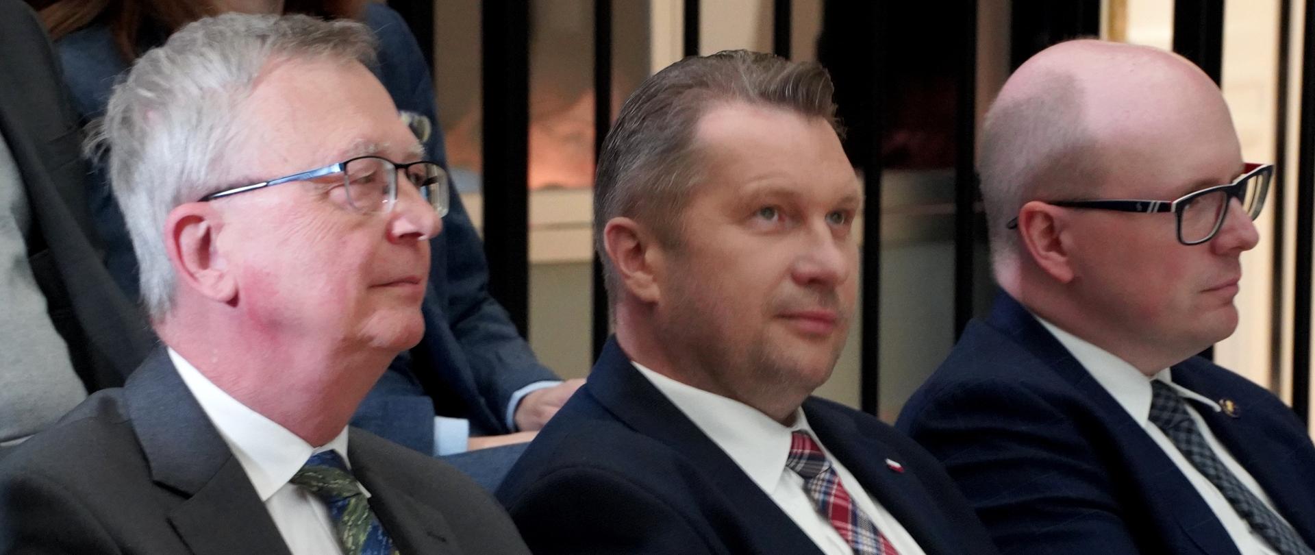 Minister Czarnek i dwóch mężczyzn w garniturach siedzą na krzesłach, za nimi inni ludzie.