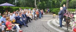 Na wyłożonym szarą kostką placu na ustawionych w rzędy krzesłach siedzą ludzie, przed nimi dwóch mężczyzn w garniturach stoi przy mikrofonach.