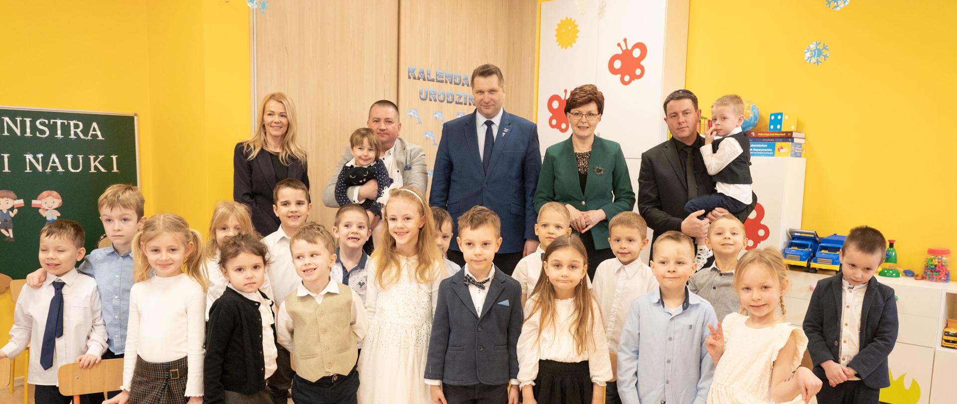 Zdjęcie zbiorowe, w sali z żółtymi ścianami w rzędzie stoją małe dzieci, za nimi kilkoro dorosłych, minister Czarnek pośrodku.